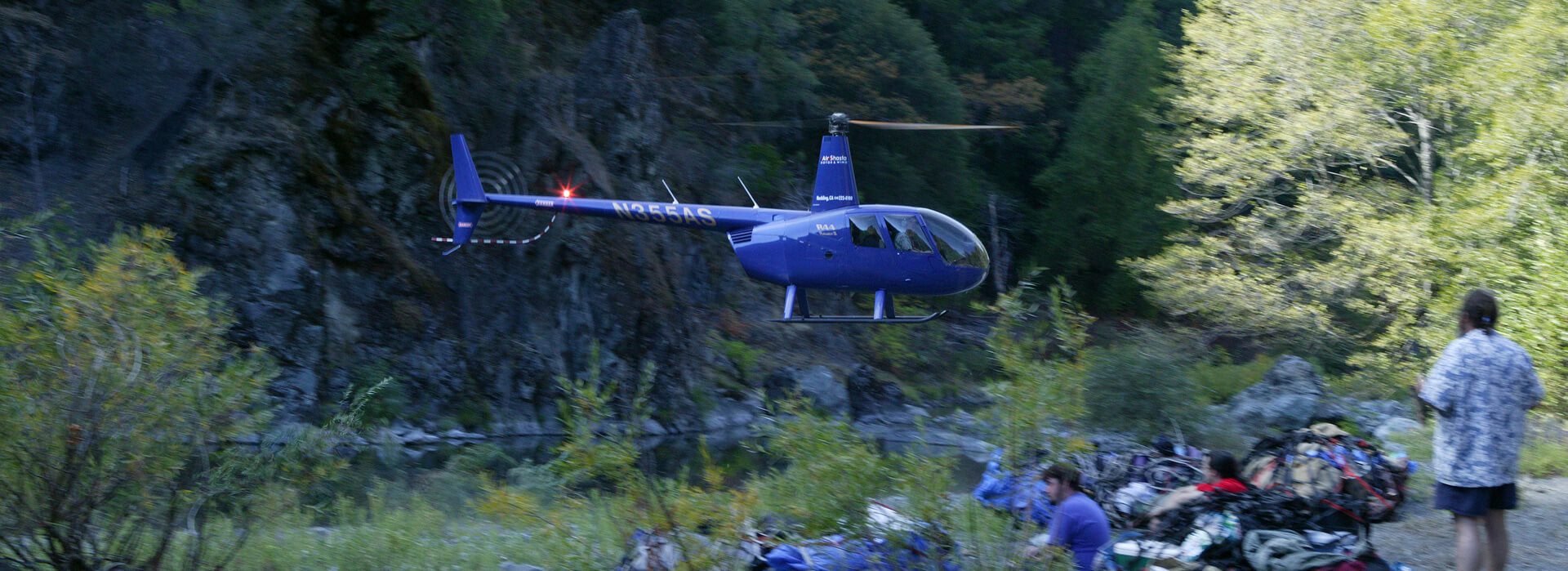 Helicopter hovers above river as jade hunters recline amongst their piled excursion gear in remote Trinity Mountain wilderness, California.